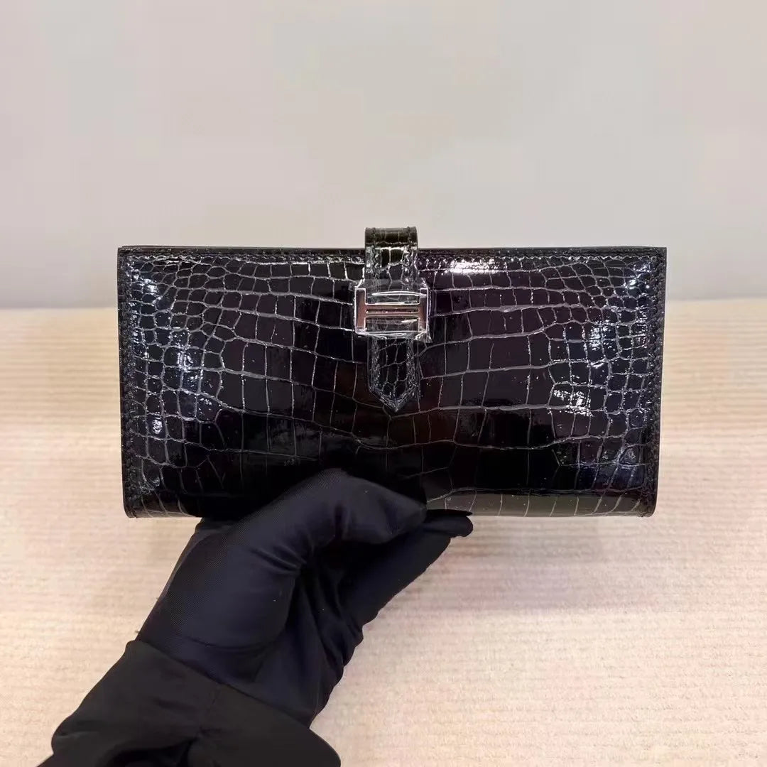 A black crocodile-embossed clutch bag with a silver-tone turn-lock clasp, held by a hand wearing a black glove against a neutral beige and gray background.