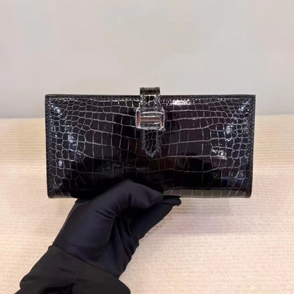 A black crocodile-embossed clutch bag with a silver-tone turn-lock clasp, held by a hand wearing a black glove against a neutral beige and gray background.
