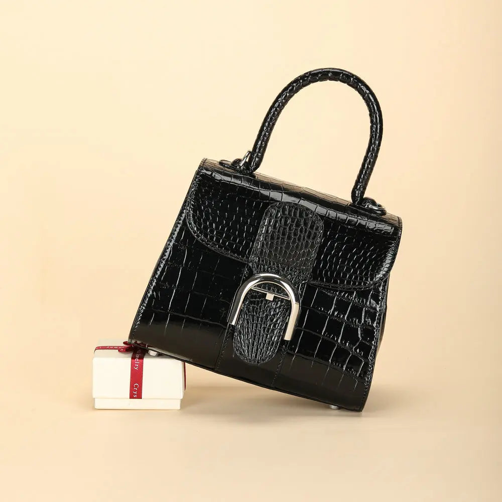 A black crocodile-embossed handbag with a silver-tone buckle closure, displayed on a beige background next to a small white gift box with a red ribbon.