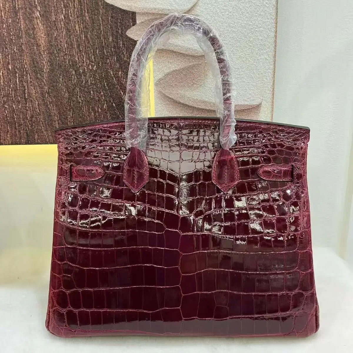 A glossy crocodile-embossed leather handbag in deep burgundy, with protective plastic wrapping on the handles, displayed against a neutral background.