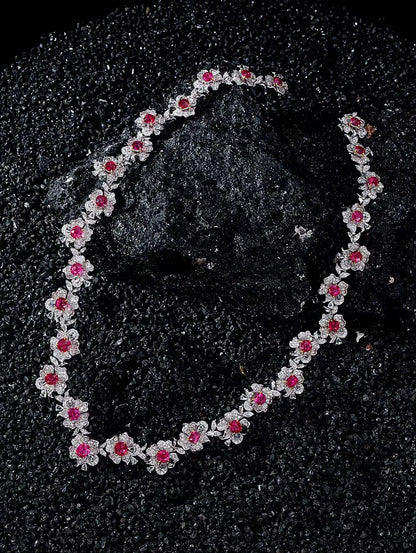 A silver-toned floral necklace with bright pink gemstone centers, arranged in an oval on a textured black background.