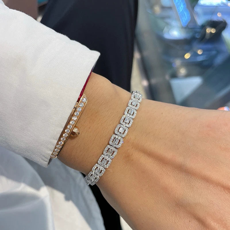 A close-up of a person's wrist wearing two bracelets: a silver-toned square-link bracelet paved with clear stones and a gold-toned bangle with a single clear stone, shown against a blurred background.