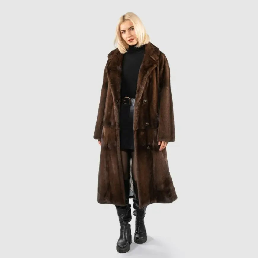 A blonde model wearing a long, dark brown fur coat over a black turtleneck, black skirt, and black leather boots, standing against a light gray studio background.