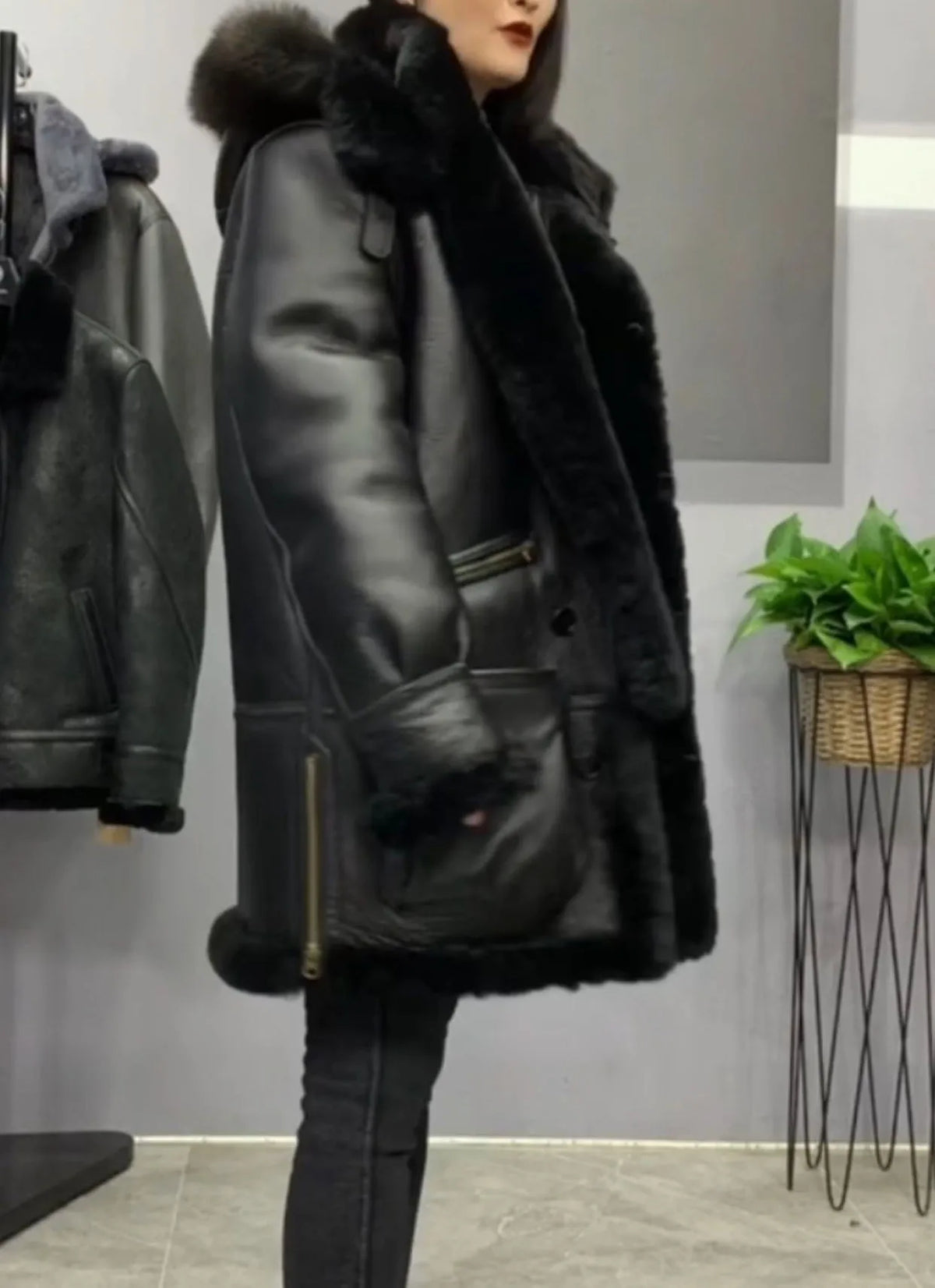 A woman wearing a black shearling-lined aviator-style jacket with a large dark fur-trimmed hood, standing in an indoor showroom. A similar jacket hangs on a rack to the left, and a potted green plant sits on a wire stand to the right.