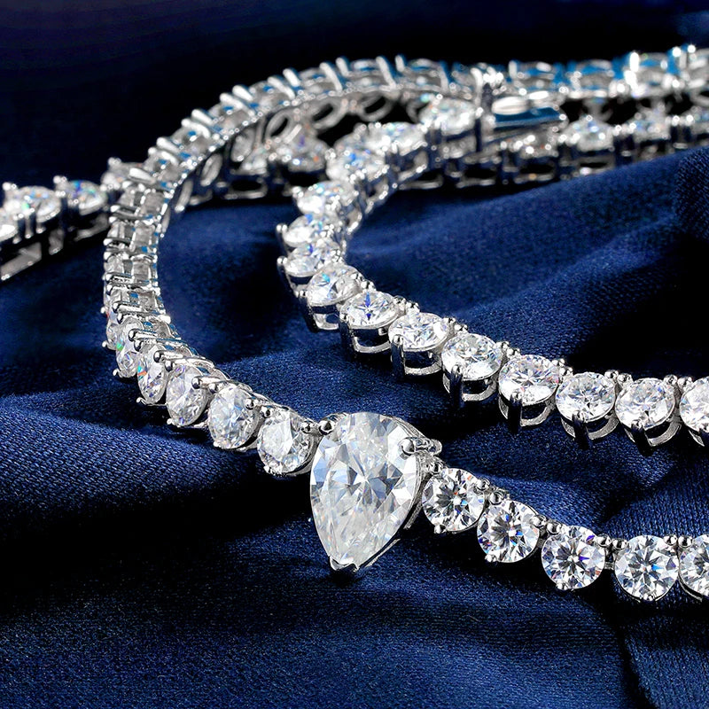 A close-up of a silver-toned tennis bracelet with a large pear-cut center stone and rows of clear round and marquise-shaped stones, displayed on dark navy blue fabric.