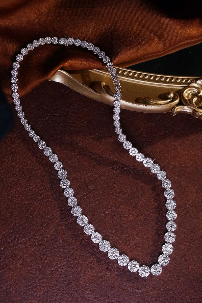 A silver tennis necklace with round-cut clear stones, arranged in a loop on a dark brown textured leather surface with a gold decorative object in the background.