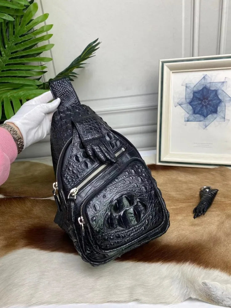 A black crocodile-embossed leather sling bag with a skull detail on the front pocket, held by a hand wearing a white glove. The bag is displayed on a brown and white cowhide rug with a green plant and a framed picture in the background.