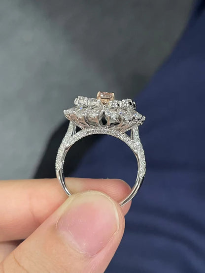 A close-up of a silver-toned ring with a cluster of clear stones and a small rose gold accent at the top, held between fingers against a dark blue and gray background.
