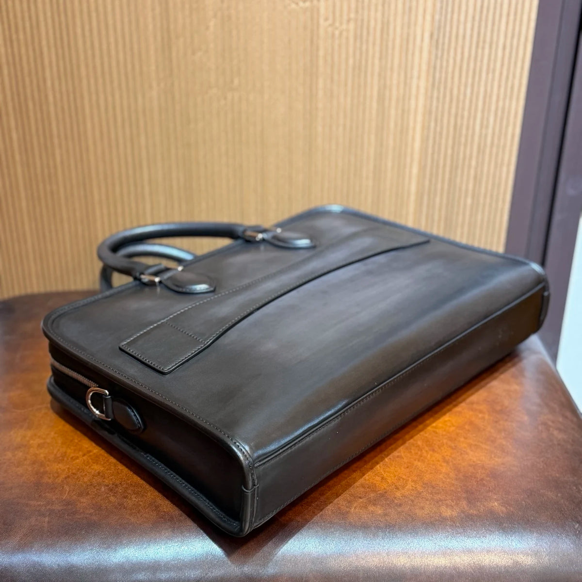A dark brown leather briefcase with a top handle, resting on a polished wooden surface against a wood-paneled background.