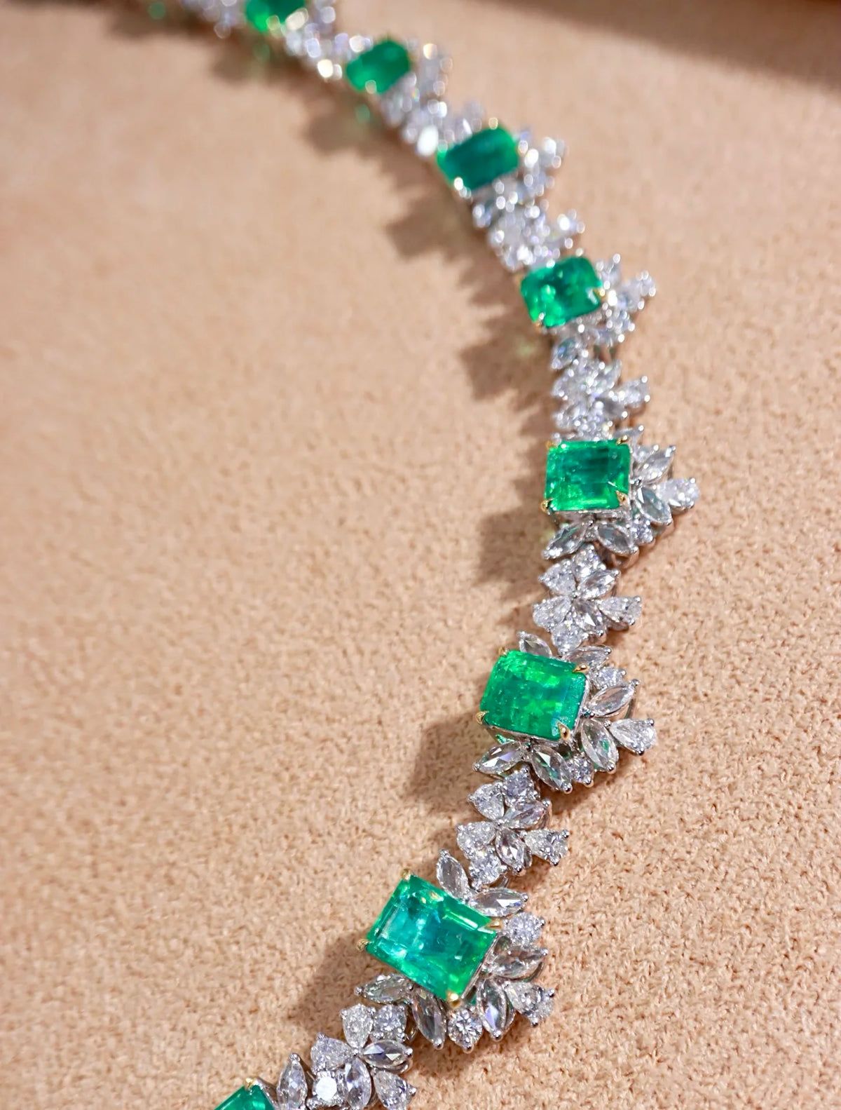 A close-up of a luxury necklace featuring vibrant green emerald-cut gemstones surrounded by sparkling clear diamond-like settings, displayed on a textured beige surface.