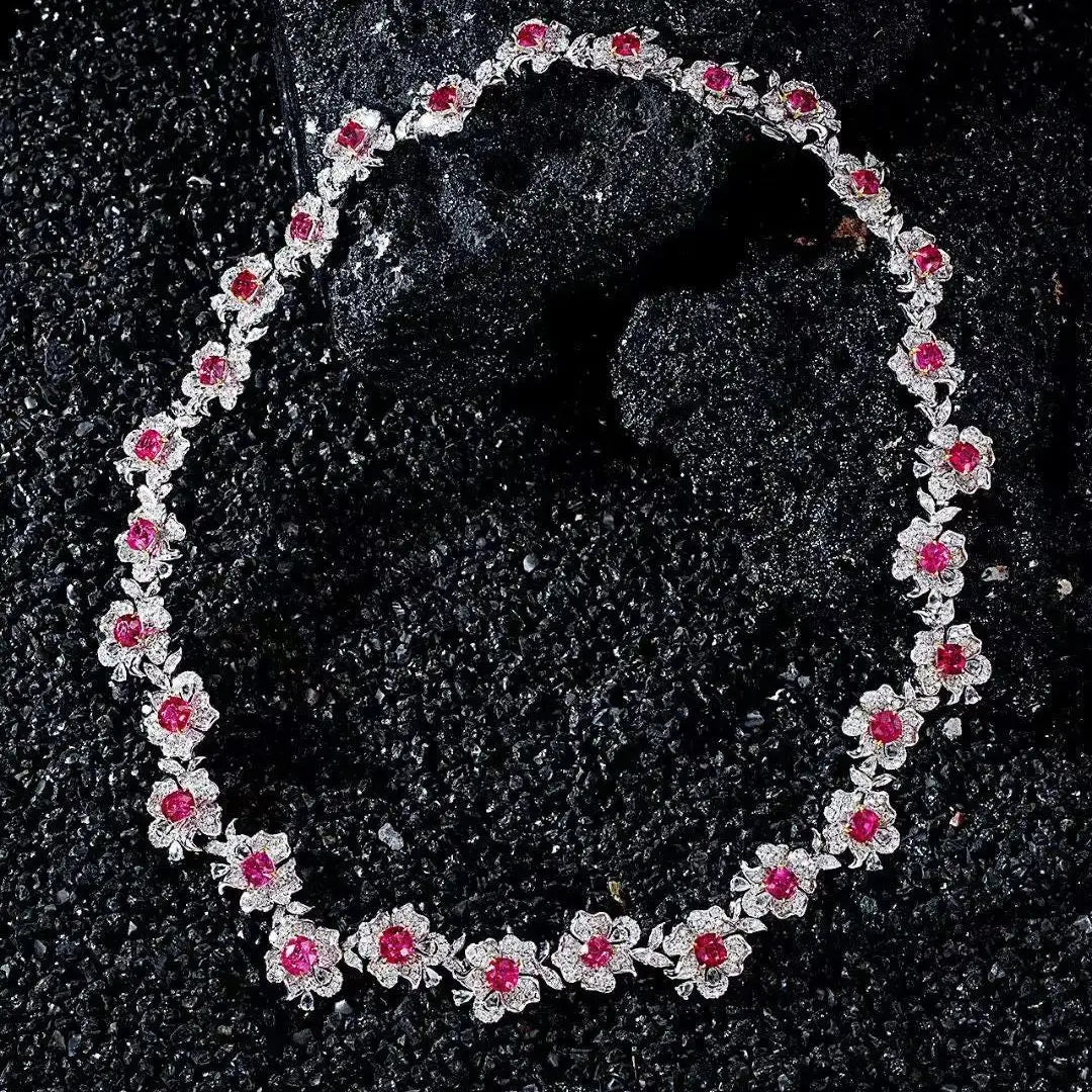 A floral necklace featuring silver-toned settings with small clear stones and bright pink gemstones, arranged in an oval on a dark textured surface.