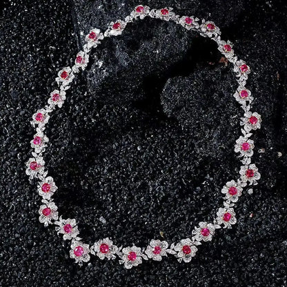 A floral necklace featuring silver-toned settings with small clear stones and bright pink gemstones, arranged in an oval on a dark textured surface.