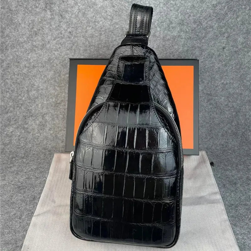 A glossy black crocodile-embossed sling bag with a top handle, standing upright on a light fabric surface in front of a gray felt backdrop and an orange box.