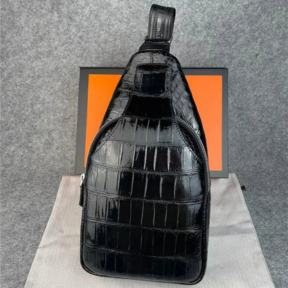 A glossy black crocodile-embossed sling bag with a top handle, standing upright on a light fabric surface in front of a gray felt backdrop and an orange box.