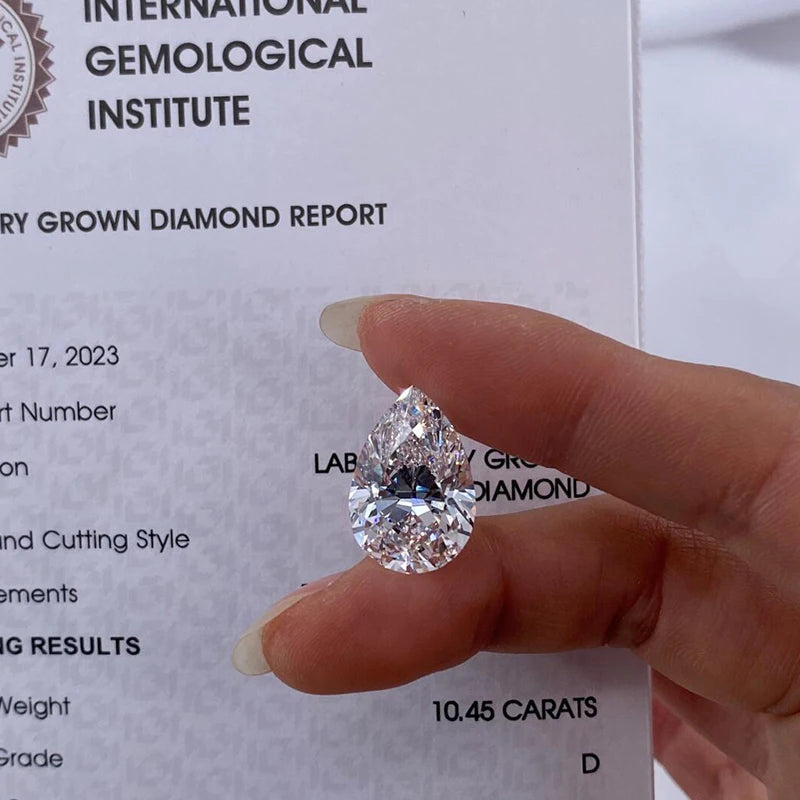 A pear-shaped brilliant-cut diamond held between fingers in front of a gemological institute report card. The certificate shows the International Gemological Institute branding and details for a 10.