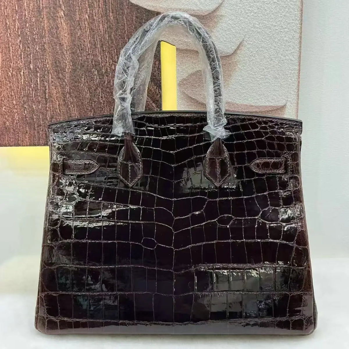 A dark brown glossy crocodile-embossed handbag with protective plastic wrapping on the handles, set against a split wood and textured wall background.