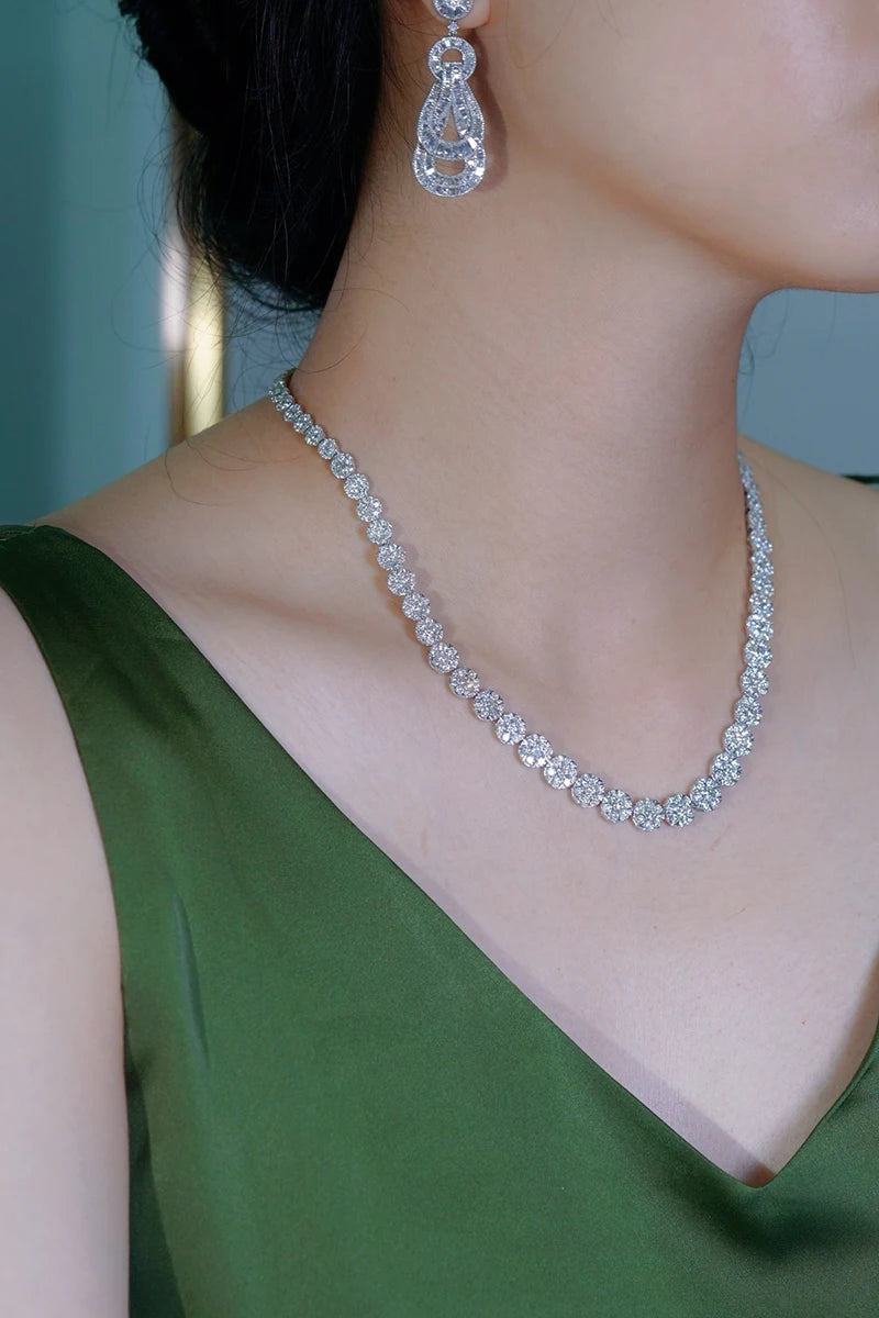 A close-up of a woman wearing a sparkling diamond tennis necklace and matching teardrop drop earrings, paired with an olive green satin top.