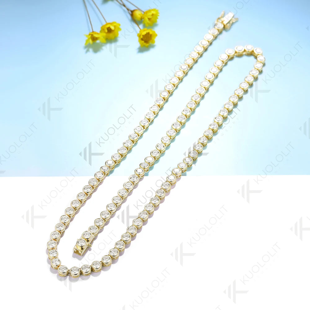 A gold-toned tennis chain necklace paved with clear round stones, arranged in an S-shape on a split light blue and white background with small yellow flowers in the top corner.