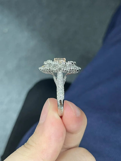 A close-up of a silver-toned ring with a large floral cluster setting and a rectangular center stone, held between fingers against a dark gray and blue background.