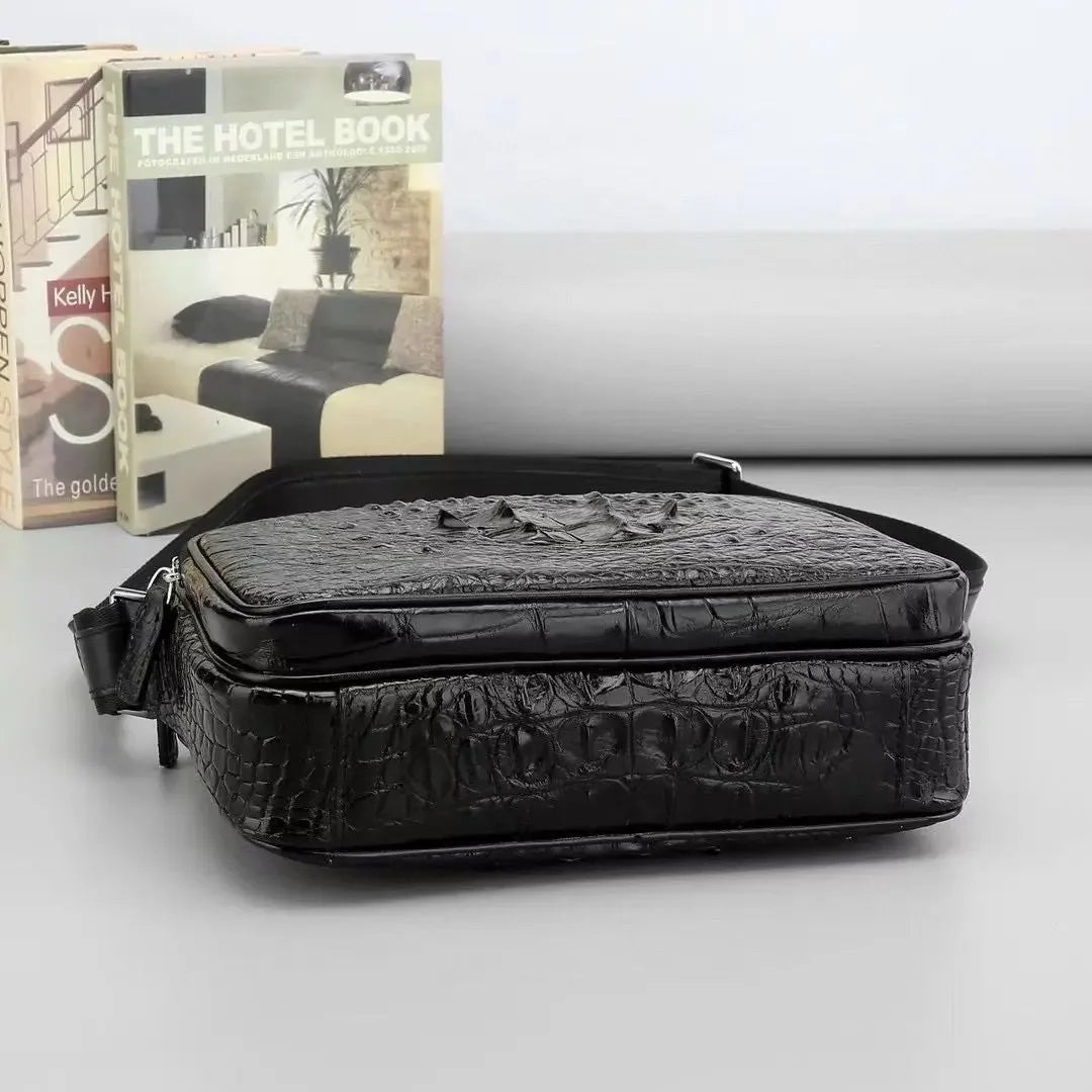 A black crocodile-embossed leather crossbody bag with a zippered top, displayed on a light gray surface with books in the background.