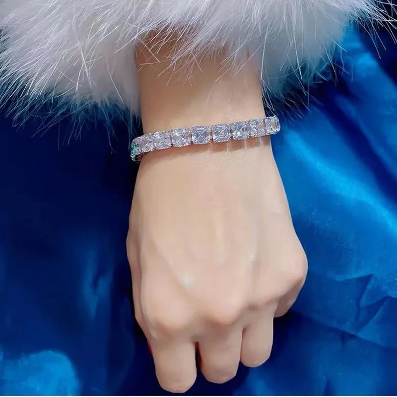 A close-up of a person's wrist wearing a sparkling silver-toned tennis bracelet with clear stones, set against a blue satin fabric and white faux fur.