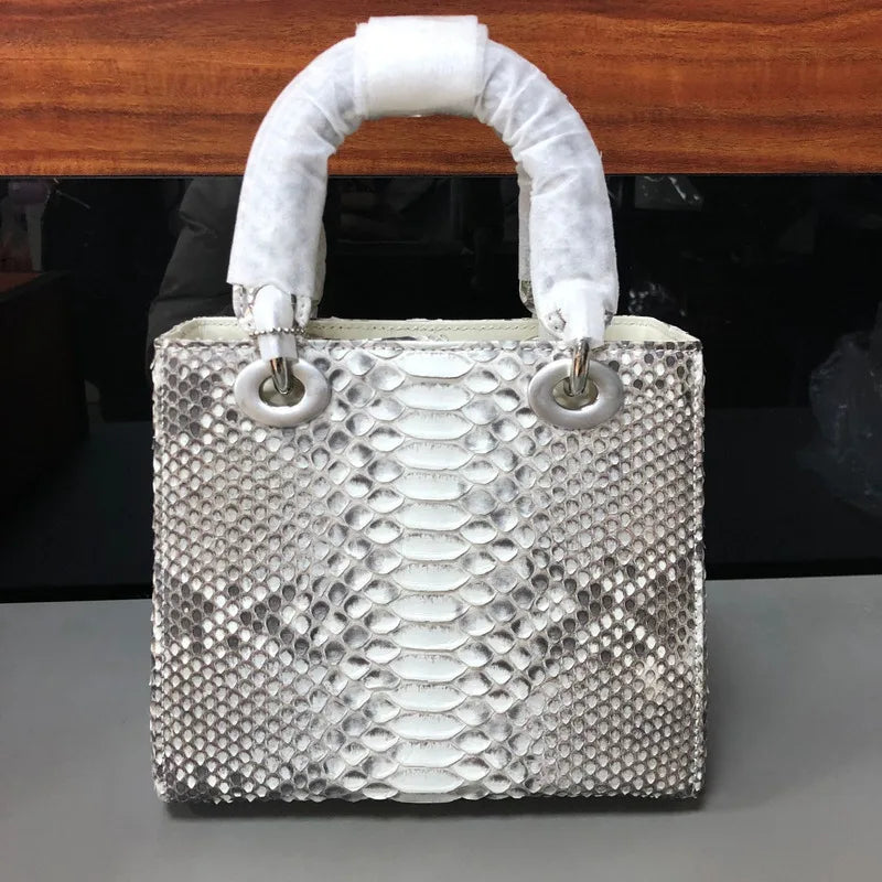 A silver snakeskin-patterned handbag with a white protective paper wrap on its top handles, resting on a gray surface in front of a dark background and wooden ledge.
