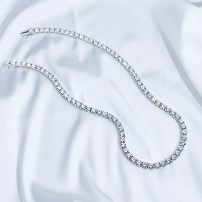 A silver tennis necklace paved with clear cubic zirconia stones, arranged in a curved S-shape on a white satin fabric background.