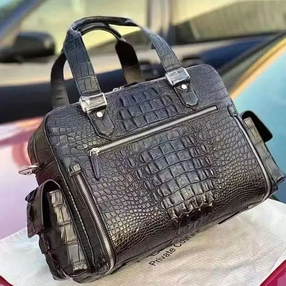 A black crocodile-embossed leather handbag with dual top handles, silver-tone hardware, and multiple zippered compartments, resting on a white dust bag in front of a blurred car background.