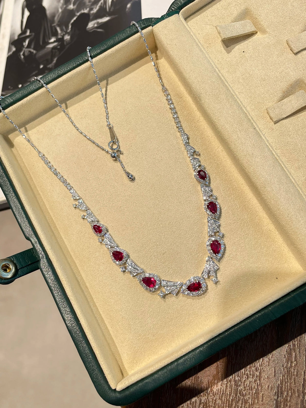 A silver-toned necklace with multiple oval red gemstones and clear stones, displayed inside an open dark green jewelry box with a beige insert.
