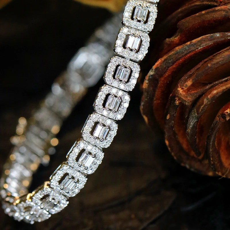 A close-up of a diamond bracelet featuring rectangular baguette-cut clear stones in silver-toned settings, arranged diagonally against a dark brown textured background.