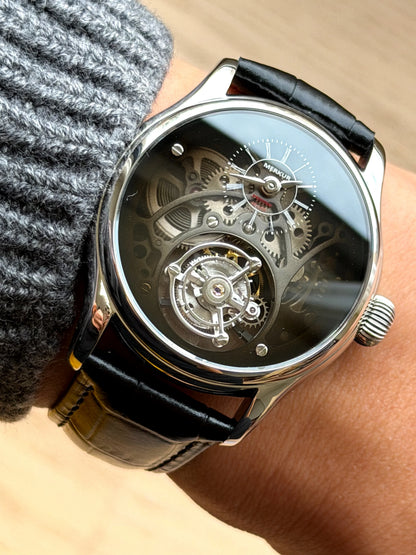 A close-up of a skeleton mechanical watch with a black leather strap worn on a wrist, showing the intricate silver-toned inner mechanism through a dark dial.
