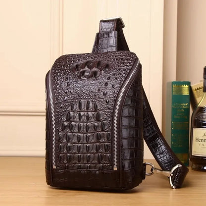 A dark brown crocodile-embossed leather sling bag with a shoulder strap, standing on a wooden surface next to a bottle and books.