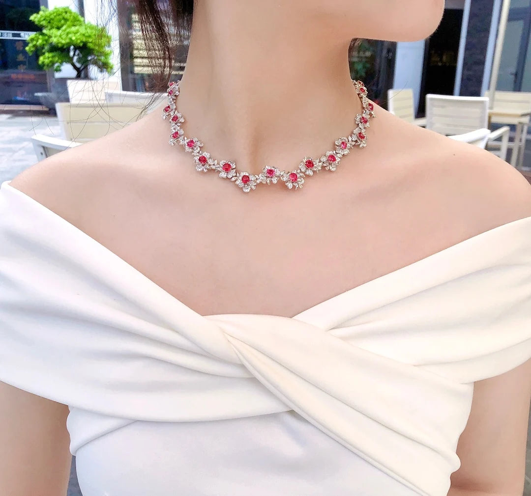 A close-up lifestyle shot of a woman wearing a silver-toned floral necklace with red gemstones, paired with a white off-the-shoulder top.