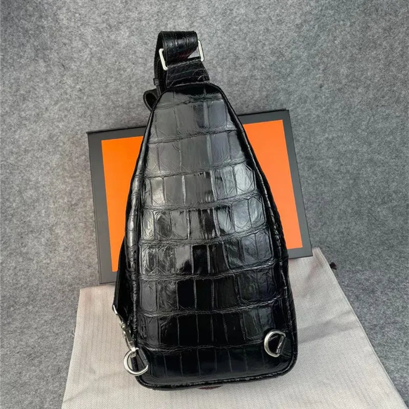 A glossy black crocodile-embossed shoulder bag with silver hardware, displayed on a light fabric surface with an orange and black box behind it against a gray felt background.