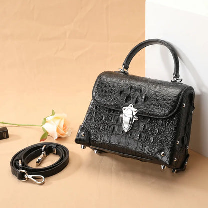 Black crocodile-embossed handbag with a top handle, silver clasp, and matching detachable shoulder strap, styled on a beige backdrop with a rose prop.