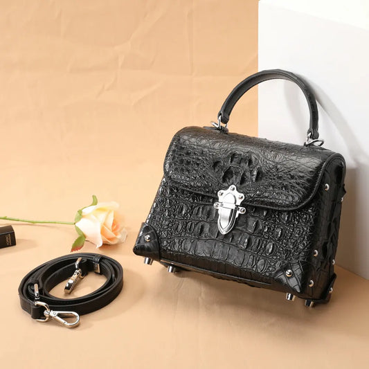 Black crocodile-embossed handbag with a top handle, silver clasp, and matching detachable shoulder strap, styled on a beige backdrop with a rose prop.