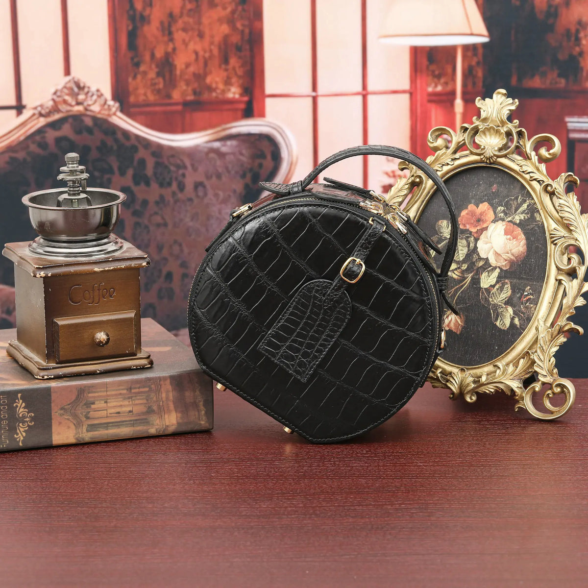 A round black faux-crocodile leather handbag with a top handle and gold-tone hardware, styled on a wooden table next to a vintage coffee grinder, a book, and an ornate gold picture frame.