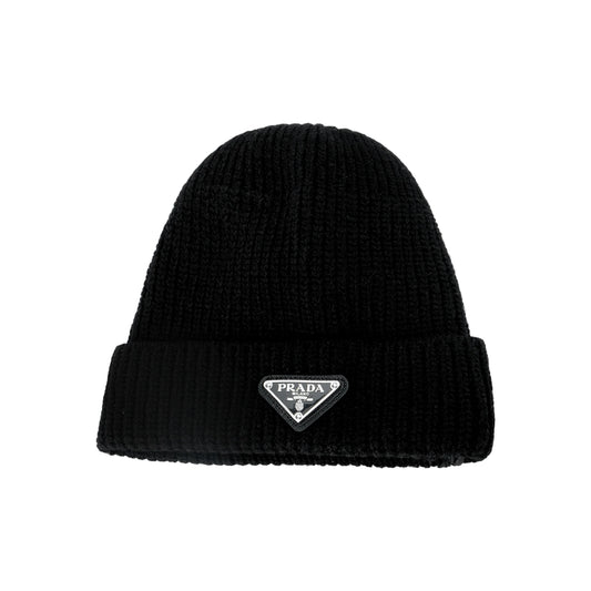 A black ribbed knit beanie hat featuring a small silver-tone triangular Prada logo patch on the front folded cuff, set against a plain white background.