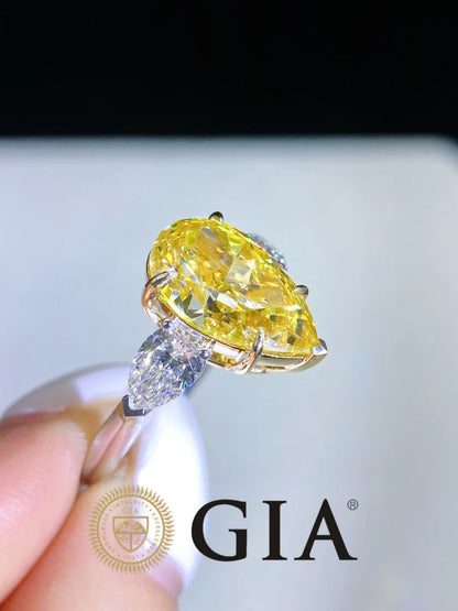 A yellow marquise-cut diamond ring held between fingers, featuring a yellow center stone and a marquise-cut diamond accent on a gold-toned band, with a GIA logo watermark at the bottom.