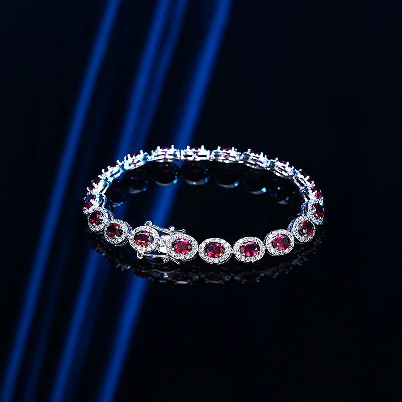A silver-toned tennis bracelet featuring round red gemstones surrounded by small clear stones, displayed on a reflective black surface with blue light streaks in the background.