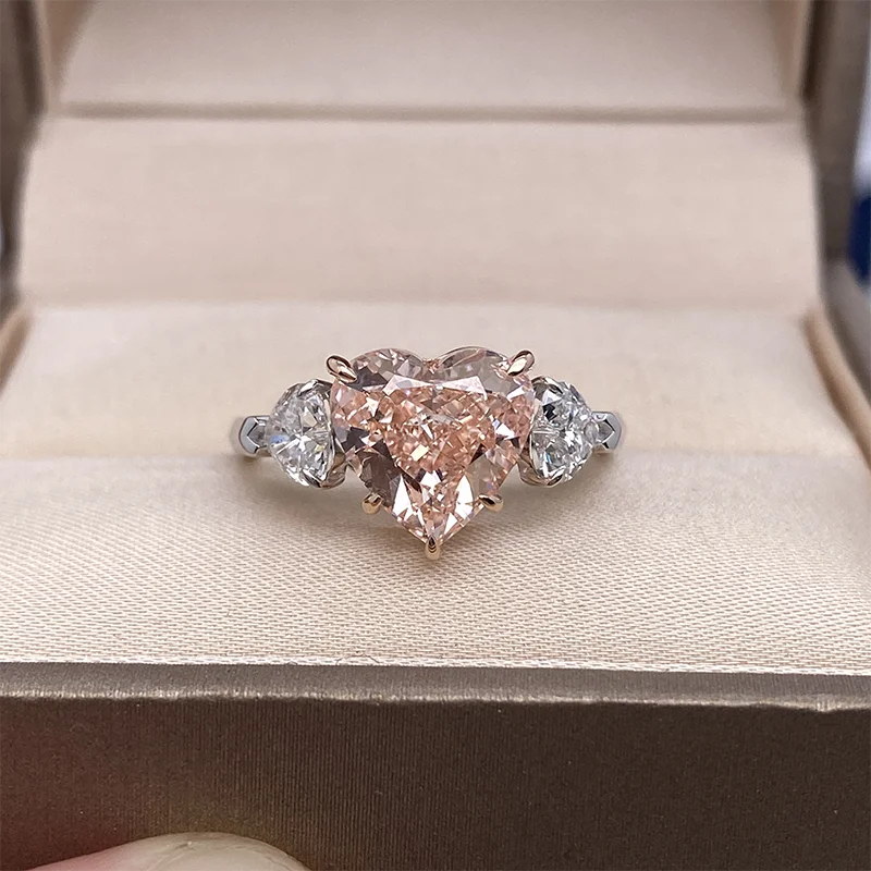 A heart-shaped diamond ring with pear-cut side diamonds set in a rose gold band, resting on a beige velvet-lined jewelry box.