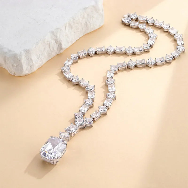 A silver-toned necklace with a row of clear rectangular crystals and a large clear rectangular pendant, arranged on a warm beige surface with a white stone prop in the corner.