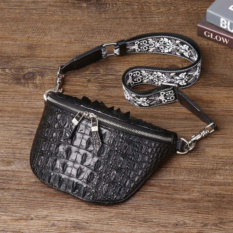 Black crocodile-embossed faux-leather crossbody bag with a patterned strap, resting on a wooden tabletop next to a book.