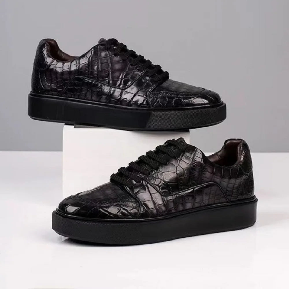 A pair of black low-top sneakers with a glossy crocodile-embossed texture, displayed on a white pedestal and a white surface against a light gray background.