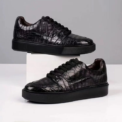 A pair of black low-top sneakers with a glossy crocodile-embossed texture, displayed on a white pedestal and a white surface against a light gray background.