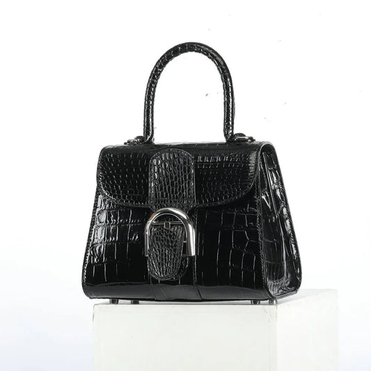 A black crocodile-embossed handbag with a structured top handle and a silver-tone metal clasp, displayed on a white block against a white background.
