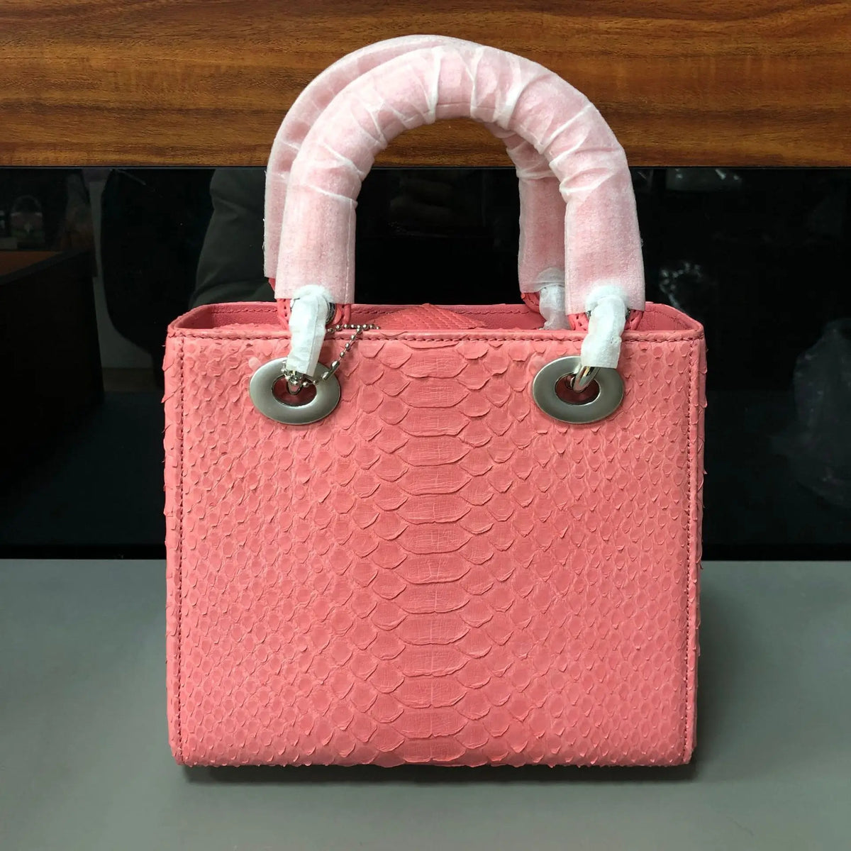 A pink textured handbag with a snakeskin pattern, featuring two rolled top handles wrapped in white protective material and silver-tone hardware, set against a dark background.