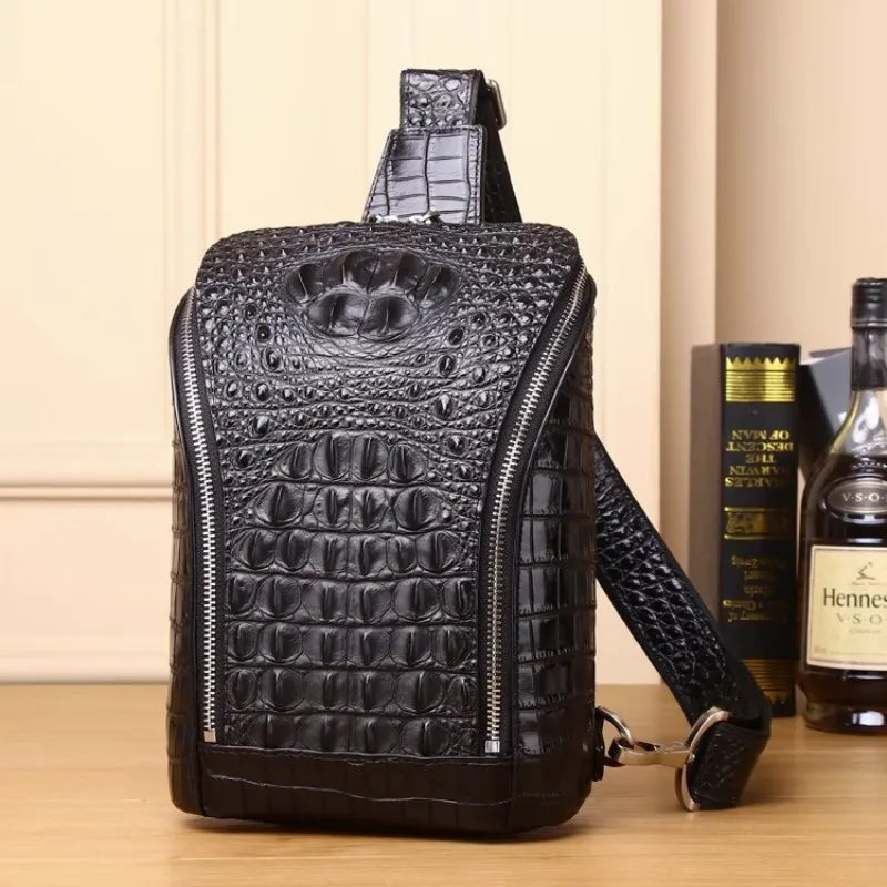 Black crocodile-embossed leather crossbody bag with a front zip pocket and shoulder strap, displayed on a wooden surface next to books and a bottle.