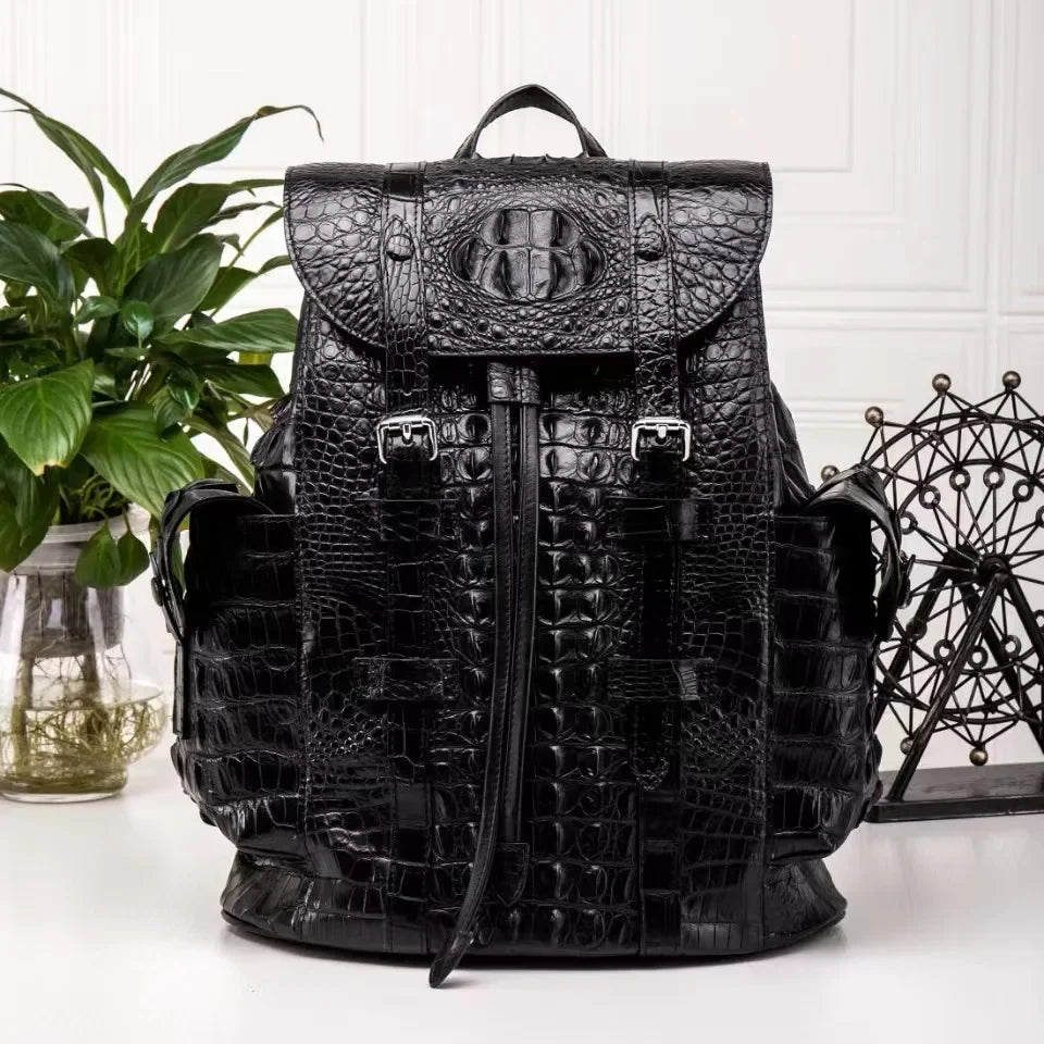 A black crocodile-embossed leather backpack with a top handle, front flap pocket, and buckle details, displayed on a white surface with a green plant and decorative metal sculpture in the background.