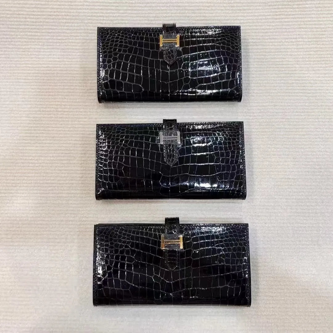 Three black crocodile-embossed leather wallet clutches are lined up vertically on a textured off-white background, each featuring a gold-tone H-shaped clasp.